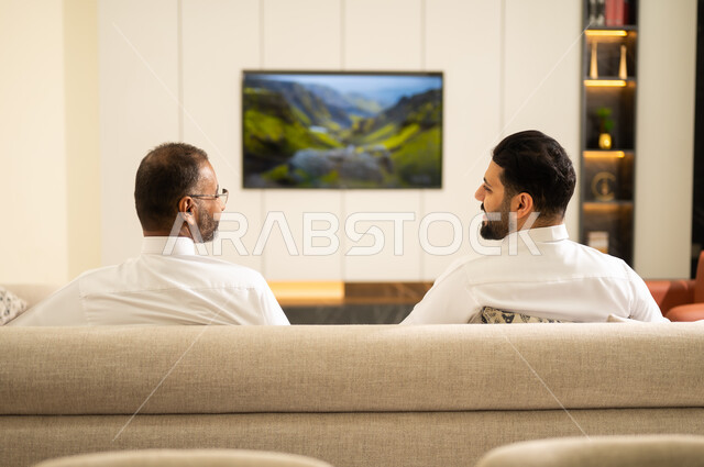 Watching the match and excitement with friends, passion for recreational activities, gestures of victory and triumph, matches and sports. A back-to-back image of two Saudi Arabian Gulf friends wearing traditional dress sitting on a comfortable couch, looking at each other with happy expressions, watching a football match.