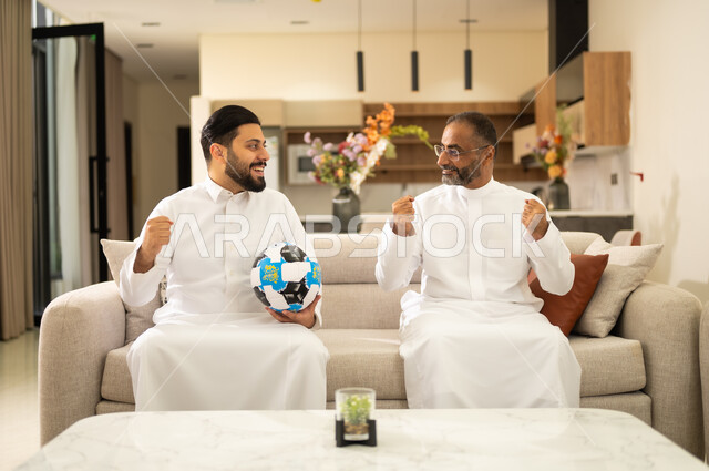 الشغف بمشاهدة الفريق المفضل , ايماءات الفرح والحماس بالانتصار ، التفاعل مع المنتخب ،المباريات والألعاب الرياضية ، صديقان عربيان خليجيان سعوديان يرفعان قبضة يدهما بتعابير الفوز، احدهما يمسك الكره بيده ، قضاء وقت ممتع بمشاهدة ومتابعة المباراة .