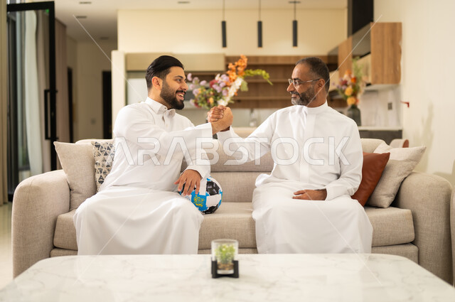 ايماءات الفرح والحماس بالانتصار , قضاء وقت ممتع بمشاهدة ومتابعة مباراة كرة القدم ، التفاعل مع المنتخب ، المباريات والألعاب الرياضية ، صديقان عربيان خليجيان سعوديان يصافحان الايدى بتعابير الفوز، احدهما يمسك الكره بيده ، الشغف بمشاهدة الفريق المفضل .