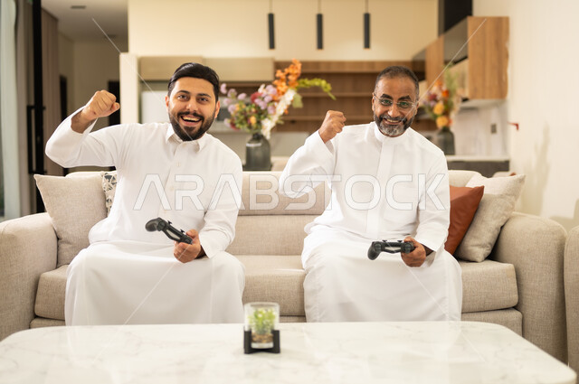 إيماءات التحدي والمتعة والفوز ، رجلان عربيان خليجيان سعوديان يمسكان عصا التحكم بتعابير السرور والسعادة، فعاليات عطلة نهاية الأسبوع ، تقنيات الكترونية حديثة ترفيهيه ، التفاعل والاندماج مع ألعاب الفيديو .