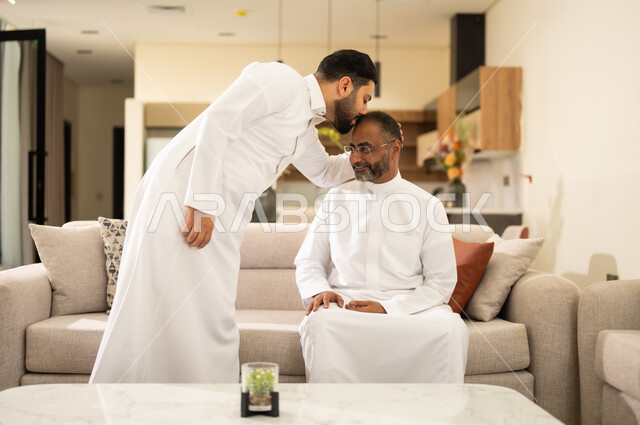 Spending quality time with family members, raising children properly, bonding and cohesion in Saudi families, a Saudi Arabian Gulf man wearing traditional clothing kissing his father's head, exchanging love and affection between parents and children.