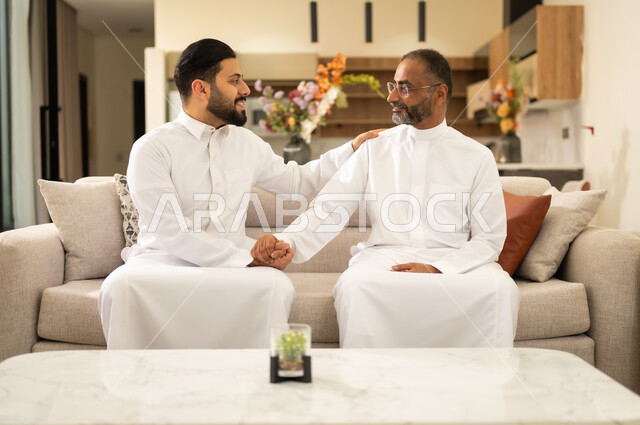 مفهوم الإحسان الى الوالدين ، الترابط والتماسك في الأسر السعودية , أب عربي خليجي سعودي يجلس على اريكة مريحة مع ابنه , النظر بإيماءات الفرح و البهجة, تبادل اطراف الحديث بين افراد الاسرة ، التربية الصالحة للأبناء .