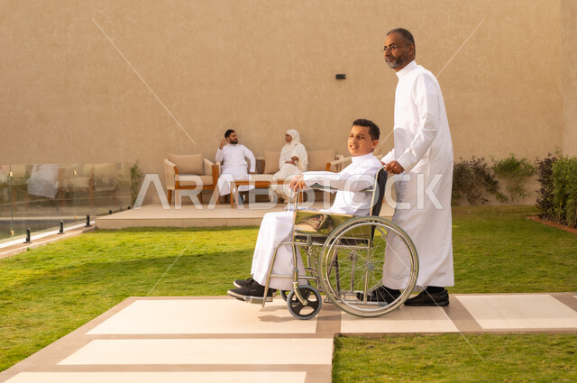 دعم المجتمع لذوي الاحتياجات الخاصه ، الاهتمام بطفل من ذوى الهمم ،  مساعدة مرضى ذوي الإعاقة الحركية على التنقل بسهولة ، قضاء الوقت في المنتزه مع العائلة ، صورة من الجانب لولد عربي خليجي سعودي مقعد يجلس على كرسي متحرك ، الاخلاص والعطف بين افراد الاسرة ، خلفية الطبيعة.