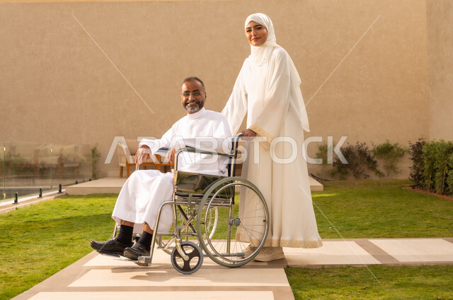 تأمين أدوات ومعدات لمساعدة المرضي ، شعور ذوي الهمم بالراحه والطمأنينه ، رجل مقعد يجلس على كرسي متحرك، امرأة عربية خليجية سعودية ترتدي العباءة والحجاب، تساعد زوجها على التنقل والحركة  ،  النظر الى الكاميرا  بايماءات الفرح والسرور ، خلفية الطبيعة.