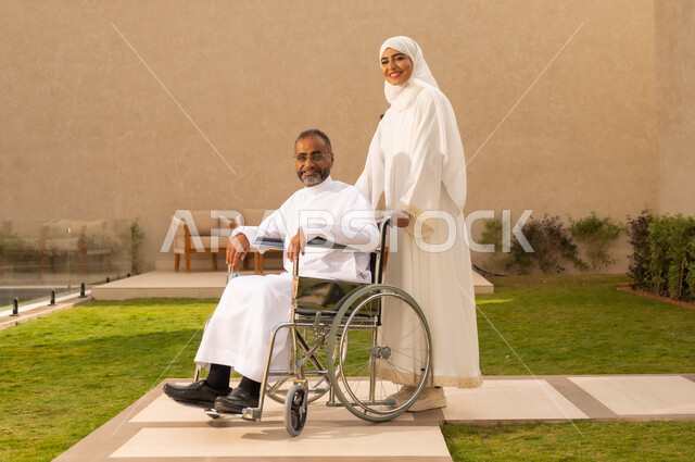 الرعاية الصحية والاعتناء باصحاب الهمم  ، تأمين أدوات ومعدات لمساعدة المرضي ، رجل مقعد يجلس على كرسي متحرك، امرأة عربية خليجية سعودية ترتدي العباءة والحجاب، تنظر الى الكاميرا  بايماءات السعادة ، تساعد زوجها على التنقل والحركة  ، خلفية الطبيعة.