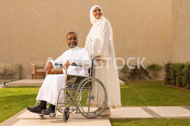 شعور ذوي الهمم بالراحه في بيئتهم ، تأمين أدوات ومعدات لمساعدة المرضي ، رجل مقعد يجلس على كرسي متحرك، امرأة عربية خليجية سعودية ترتدي العباءة والحجاب، تنظر الى شئ ما بايماءات السعادة ، تساعد زوجها على التنقل والحركة  ، خلفية الطبيعة.