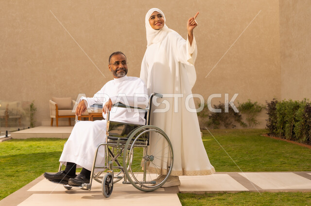 الاخلاص والعطف بين الزوجين ،جودة حياة ذوي الهمم في السعودية ، تأمين أدوات ومعدات لمساعدة المرضي ، رجل مقعد يجلس على كرسي متحرك، امرأة عربية خليجية سعودية ترتدي العباءة والحجاب، تشير الى شئ ما بايماءات السعادة ، تساعد زوجها على التنقل والحركة ،  قضاء الوقت في المنتزه مع العائلة ، خلفية الطبيعة.