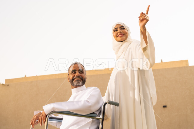 التحسين من نفسية اصحاب الهمم ، نمط حياة ذوي الإعاقة الحركية ، تأمين أدوات ومعدات لمساعدة المرضي ، رجل مقعد يجلس على كرسي متحرك، امرأة عربية خليجية سعودية ترتدي العباءة والحجاب، تشير الى شئ ما بايماءات السعادة ، تساعد زوجها على التنقل والحركة ،  قضاء الوقت في المنتزه مع العائلة ، خلفية الطبيعة.