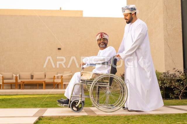 النظر الى شئ ما بايماءات السعادة ، نمط حياة ذوي الإعاقة الحركية ، تأمين أدوات ومعدات لمساعدة المرضي ، رجل مقعد يجلس على كرسي متحرك، صورة من الجانب لشاب عربي خليجي عماني يرتدي الدشداشة والكمة، يساعد والده على التنقل والحركة ،  قضاء الوقت في المنتزه مع العائلة ، خلفية الطبيعة.