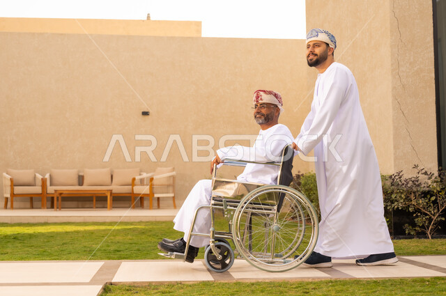 العطف والحب بين أفراد الأسرة, العنايه بأصحاب الهمم وتأمين احتياجاتهم ، نمط حياة ذوي الإعاقة الحركية ، تأمين أدوات ومعدات لمساعدة المرضي ، رجل مقعد يجلس على كرسي متحرك، صورة من الجانب لشاب عربي خليجي عماني يرتدي الدشداشة والكمة، يساعد والده على التنقل والحركة ،  قضاء الوقت في المنتزه مع العائلة .