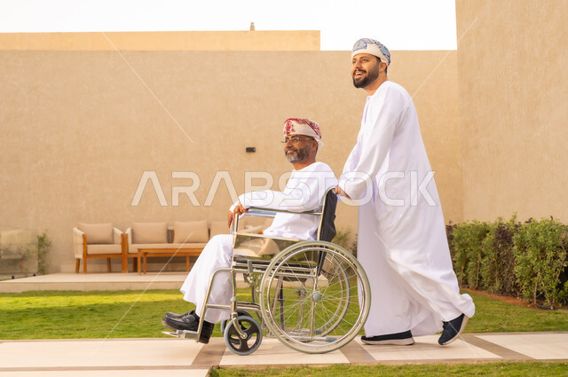 إيماءات البشاشه والمعاملة الحسنة, العنايه بأصحاب الهمم وتأمين احتياجاتهم ، نمط حياة ذوي الإعاقة الحركية ، تأمين أدوات ومعدات لمساعدة المرضي ، رجل مقعد يجلس على كرسي متحرك، صورة من الجانب لشاب عربي خليجي عماني يرتدي الدشداشة والكمة، يساعد والده على التنقل والحركة ،  قضاء الوقت في المنتزه مع العائلة .