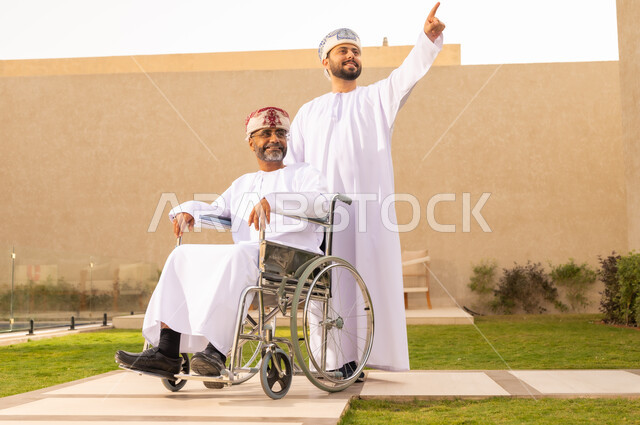 العنايه بأصحاب الهمم وتأمين احتياجاتهم ، إيماءات السعادة والسرور بالخروج فى الهواء الطلق ، نمط حياة ذوي الإعاقة الحركية ، تأمين أدوات ومعدات لمساعدة المرضي ، رجل مقعد يجلس على كرسي متحرك، شاب عربي خليجي عماني يرتدي الدشداشة والكمة و يشير الى شئ ما ، يساعد والده على التنقل والحركة ،  قضاء الوقت في المنتزه مع العائلة .