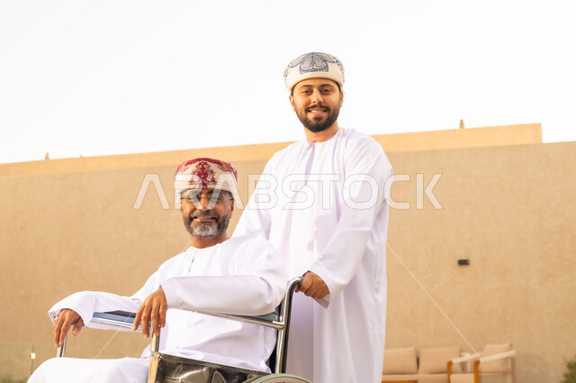 نمط حياة ذوي الإعاقة الحركية ، النظر الى الكاميرا بإيماءات السعادة والسرور ، تأمين أدوات ومعدات لمساعدة ذوى الهمم ، رجل مقعد يجلس على كرسي متحرك، شاب عربي خليجي عماني يرتدي الدشداشة والكمة ، يساعد والده على التنقل والحركة،  قضاء الوقت في المنتزه مع العائلة .