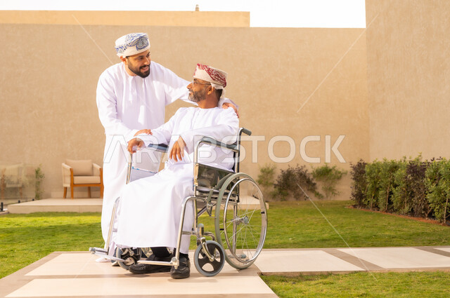 أسلوب حياة أصحاب الهمم، إيماءات السعادة والسرور بالخروج في الهواء الطلق ، تأمين أدوات ومعدات تساعد ذوى الهمم ، نمط حياة ذوي الإعاقة الحركية ، رجل مقعد يجلس على كرسي متحرك، شاب عربي خليجي عماني يرتدي الدشداشة والكمة ، يساعد والده على التنقل والحركة،  قضاء الوقت في المنتزه مع العائلة .