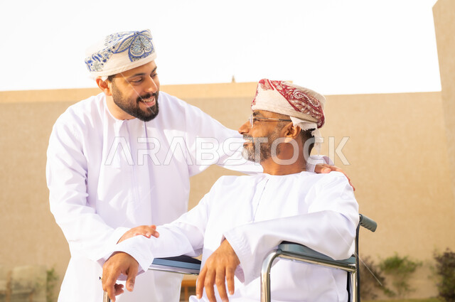 مفهوم الرعاية والعناية بذوى الهمم ، تأمين أدوات ومعدات تساعد المرضى على التنقل والحركة، نمط حياة ذوي الإعاقة الحركية  ، رجل مقعد يجلس على كرسي متحرك، شاب عربي خليجي عماني يرتدي الدشداشة والكمة برفقة والده في الحديقة ، قضاء الوقت في المنتزه مع العائلة .