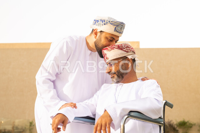 شعور ذوى الهمم بالإنتماء والتقدير ، تأمين أدوات ومعدات تساعد المرضى على التنقل والحركة، نمط حياة ذوي الإعاقة الحركية ، رجل مقعد يجلس على كرسي متحرك، صورة مقربة لشاب عربي خليجي عماني يرتدي الدشداشة والكمة ، يقبل والده بايماءات الاحترام ،  قضاء الوقت في المنتزه مع العائلة .