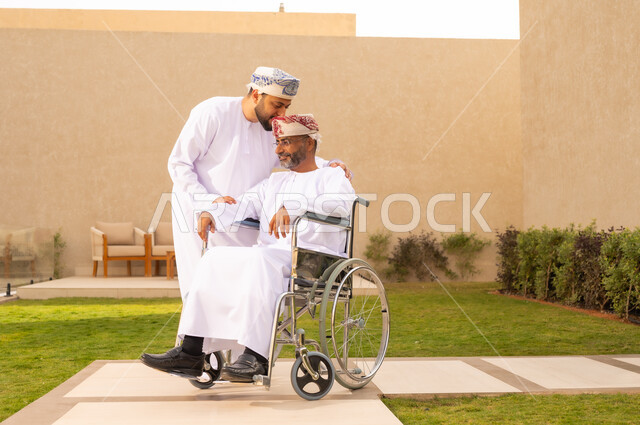 دعم الابناء لوالديهم ، تأمين أدوات ومعدات تساعد المرضى على التنقل والحركة، نمط حياة ذوي الإعاقة الحركية ، رجل مقعد يجلس على كرسي متحرك، شاب عربي خليجي عماني يرتدي الدشداشة والكمة ، يقبل والده بايماءات الاحترام ،  قضاء الوقت في المنتزه مع العائلة