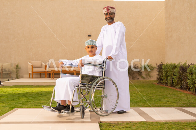 الاهتمام بطفل من ذوى الهمم ، الجلوس على كرسي متحرك وقضاء الوقت في المنتزه مع العائلة، صورة من الجانب لولد عربي خليجي عماني يرتدي الدشداشة والكمة يجلس على كرسي متحرك ,   ينظر الى الكاميرا بايماءات السعادة ، خلفية الطبيعة.