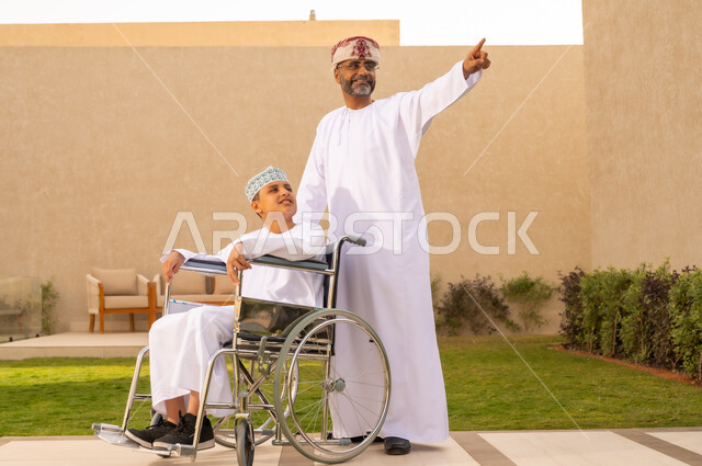 تأمين أدوات ومعدات تساعد أصحاب الهمم على التنقل والحركة، نمط حياة ذوي الإعاقة الحركية  ، ولد مقعد يجلس على كرسي متحرك، رجل عربي خليجي عماني يرتدي الدشداشة والكمة برفقة ابنه في الحديقة، يشير الى شئ ما بايماءات السعادة ، مفهوم الرعاية والعناية بالمرضى والمعاقين .