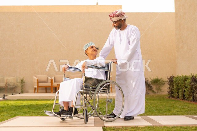 إيماءات السعادة والمعاملة الحسنة، الجلوس على كرسي متحرك وقضاء الوقت في المنتزه مع العائلة،ولد عربي خليجي عماني من ذوي الهمم يرتدي الدشداشة والكمة يجلس على كرسي متحرك , اهتمام ودعم المجتمع لذوي الاحتياجات الخاصه, خلفية الطبيعة.