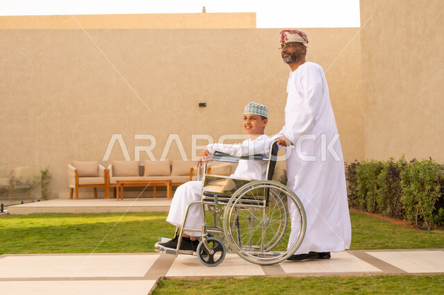 اهتمام ودعم المجتمع لذوي الاحتياجات الخاصه، الجلوس على كرسي متحرك وقضاء الوقت في المنتزه مع العائلة، صورة من الجانب لولد عربي خليجي عماني من ذوي الهمم يرتدي الدشداشة والكمة يجلس على كرسي متحرك ,  خلفية الطبيعة.