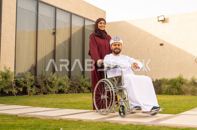 تأمين احتياجات أصحاب الهمم ، الإخلاص والعطف بين الأزواج ، مساعدة مرضى ذوي الإعاقة الحركية على التنقل بسهولة, امرأة عربية خليجية عمانية ترتدي العباءة والحجاب برفقة زوجها في إحدى الحدائق، رجل عماني مقعد يجلس على كرسي متحرك ، ينظر الى الكاميرا بايماءات السعادة .