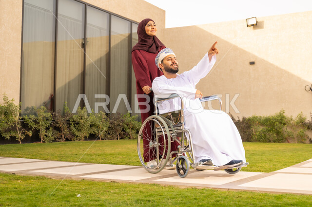 نمط حياة أصحاب الهمم ، مساعدة مرضى ذوي الإعاقة الحركية على التنقل بسهولة, الإخلاص والعطف بين الأزواج ، امرأة عربية خليجية عمانية ترتدي العباءة والحجاب برفقة زوجها في إحدى الحدائق، رجل عماني مقعد يجلس على كرسي متحرك ، يشير الى شئ ما بايماءات السعادة .