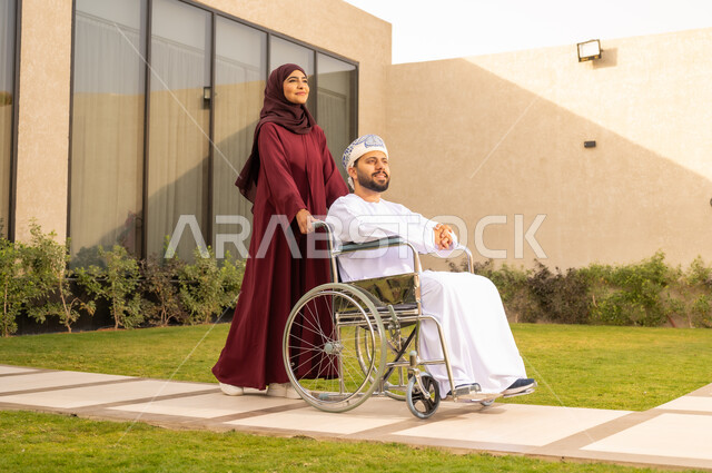 الإخلاص والعطف بين الأزواج، التحسين من نفسية أصحاب الهمم ، مساعدة مرضى ذوي الإعاقة الحركية على التنقل بسهولة, امرأة عربية خليجية عمانية ترتدي العباءة والحجاب برفقة زوجها في إحدى الحدائق، رجل عماني مقعد يجلس على كرسي متحرك .