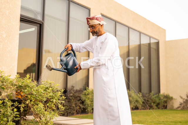 مفهوم الرفاهية والمسؤولية البيئية ، رجل عربي خليجي عماني يقوم باستخدام وعاء السقي في ري النباتات ، استخدام مرش المياة ، قضاء الوقت الممتع في إحدى الشاليهات ،مفهوم النظافة والاهتمام بحديقة المنزل ، خلفية الطبيعة .
