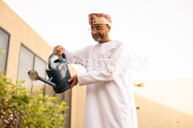 قضاء وقت ممتع فى الحديقة ، رجل عربي خليجي عماني يقوم باستخدام وعاء السقي في ري النباتات ، استخدام مرش المياة ، مفهوم النظافة والاهتمام بحديقة المنزل ، خلفية الطبيعة .
