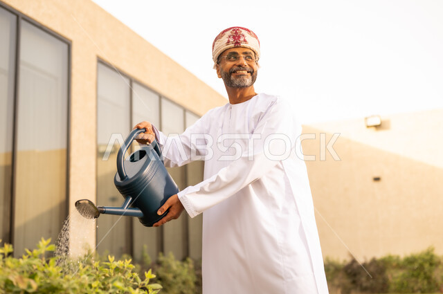 Looking at something with happy gestures, an Omani Arabian Gulf man using a water sprinkler to water plants, spending a pleasant time in a chalet, taking care of plants in the garden, a nature background.