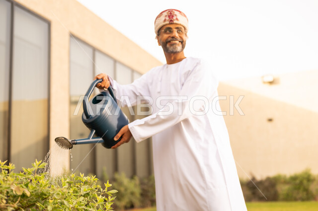 رجل عربي خليجي عماني يقوم باستخدام وعاء السقي في ري النباتات ،  اجواء الطبيعة الخلابة ، قضاء الوقت الممتع في إحدى الشاليهات ، مفهوم النظافة والاهتمام بحديقة المنزل ،  الاعتناء والاهتمام بالنباتات الخضراء .