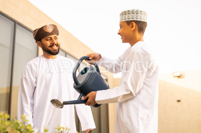 رجل عربي خليجي عماني يقضي وقت ممتع مع إبنه في ري النباتات بإستخدام وعاء السقي ، فعاليات وأجواء ترفيهية ، ايماءات السعادة على وجوههم ، خلفية الطبيعة ، الاجواء الطبيعية الجميلة ، المتعة و التسليه والترفيه ، مشاركة عائلية دافئة .