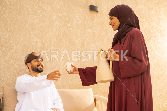 ضيافة تقليدية بايماءات الحب والأصالة ، الاستمتاع باللحظات الدافئة ، امرأة عربية خليجية عمانية تقدم الضيافة لزوجها ، روح التفاهم والمودة في الحياة الزوجية الخليجية . مفهوم الألفة بين الزوجين .