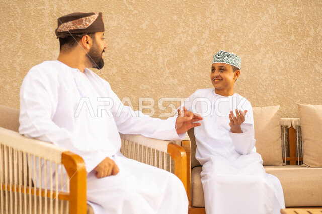 رعاية الآباء بأبنائهم والاهتمام بهم ، اب عربي خليجي عماني يرتدي الدشداشة والكمة ، يجلس مع ابنه و يتبادلان الحديث بايماءات وجه سعيدة ، اجواء عائلية دافئة ، بر الوالدين والاحسان اليهم
