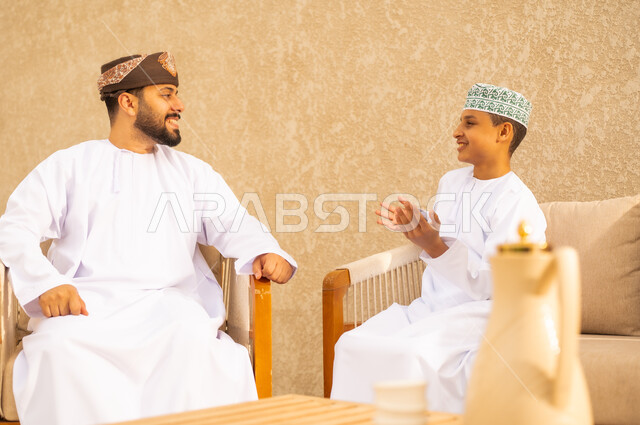 إيماءات السعادة والاستمتاع بالحديث مع الأبناء ، اب عربي خليجي عماني يرتدي الدشداشة والكمة ، يجلس مع ابنه و يتبادلان الحديث بايماءات وجه سعيدة ، اجواء عائلية دافئة ، بر الوالدين والاحسان اليهم