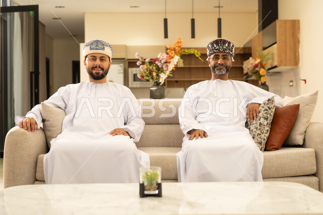 مفهوم الرابطة الأبوية, ايماءات الحب مع الأبناء, قضاء الأوقات برفقة الأبناء ، اب عربي خليجي عماني يجلس مع ابنه على كنبه مريحه في غرفه المعيشه ، اجواء عائلية دافئة بايماءات وجه سعيدة، بر الوالدين والاحسان اليهم .