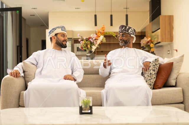 Enjoying building an intimate relationship with children, a warm family atmosphere with happy facial gestures, an Arab Gulf Omani father sitting with his son on a comfortable couch in the room Living together and exchanging conversations, the concept of honoring and being kind to parents.