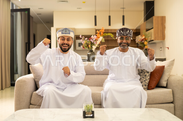 رفع قبضة اليد للاعلى بتعابير الفوز و الربح ، استخدام التكنولوجيا الحديثة، رجلان عربيان خليجيان عمانيان يجلسان على كنبه مريحه في غرفه المعيشه ، الاستمتاع والتسلية في العطلة .