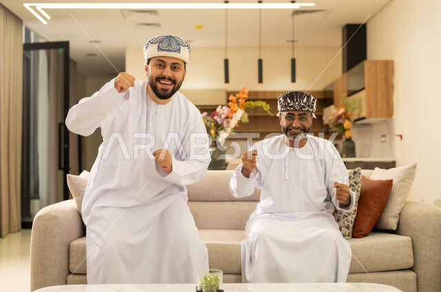 إيماءات الفوز  والانتصار فى اللعبة ، استخدام التكنولوجيا الحديثة، رجلان عربيان خليجيان عمانيان يجلسان على كنبه مريحه في غرفه المعيشه ، الاستمتاع والتسلية في العطلة .