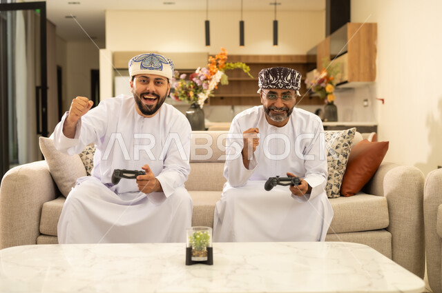 إيماءات التحدي والمتعة والتسليه، استخدام التكنولوجيا الحديثة، قضاء الاوقات السعيده باللعب مع الابناء، اب عربي خليجي عماني يجلس مع ابنه على كنبه مريحه في غرفه المعيشه ويمسكان عصا التحكم ، اللعب بألعاب الفيديو .