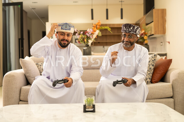 الاستمتاع والتسلية في العطلة ، استخدام التكنولوجيا الحديثة، رجلان عربيان خليجيان عمانيان يجلسان على كنبه مريحه في غرفه المعيشه يمسكان عصا التحكم ، إيماءات الفوز والخسارة فى اللعبة .