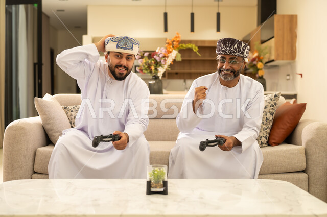 Spending happy times playing with their children: an Arab Gulf Omani father sitting with his son on a comfortable sofa in the living room, holding a joystick, playing video games, gestures of challenge, fun, and entertainment, using modern technology.