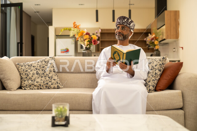 قراءة الأذكار والأدعية ، تلاوة وقراءة القرآن الكريم وتدبر معانيه، رجل عربي خليجي عماني يرتدي الدشداشة والعمامة  يحمل بيده المصحف الشريف وينظر الى شئ ما ، الإكثار من ذكر الله تعالى ، إسلاميات وعبادة ،التقرب الى الله ، الاجتهاد في العبادة .