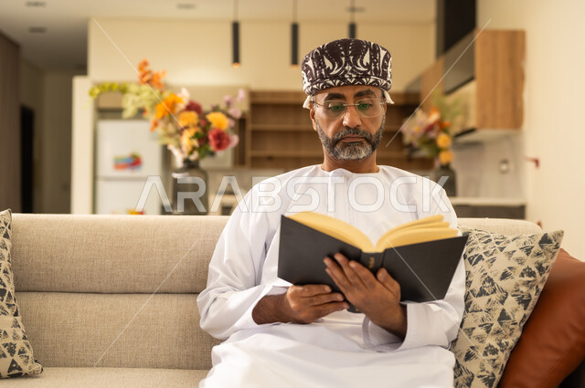 الاستمتاع بقراءة الروايات والقصص ، الشغف بممارسة الهوايات المفيدة ، صورة مقربة لرجل عربي خليجي عماني يرتدي الدشداشة والكمة يجلس على اريكة مريحة ويحمل بيده كتاب تثقيفي ، قراءة الكتب بتمعن ، الاستمتاع بقراءة الروايات والقصص .