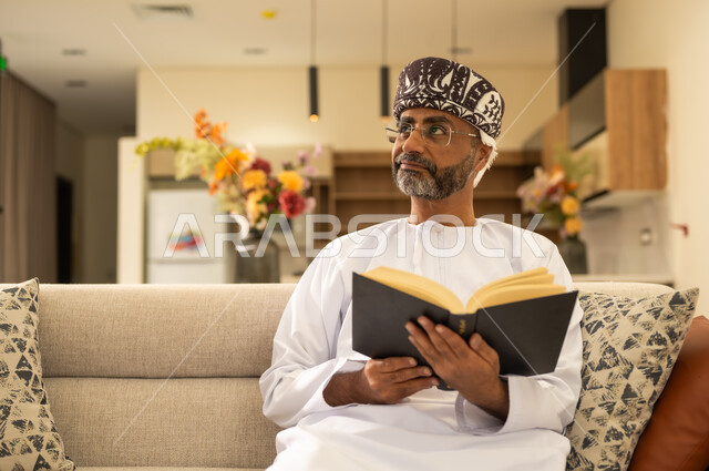 استغلال الوقت في التثقف،  ثقافة التعلم الذاتي، قراءة الكتب بتمعن ، رجل عربي خليجي عماني يرتدي الدشداشة والكمة يجلس على اريكة مريحة ويحمل بيده كتاب تثقيفي ،  النظر بعيداً الى مكان ما ، الاستمتاع بقراءة الروايات والقصص