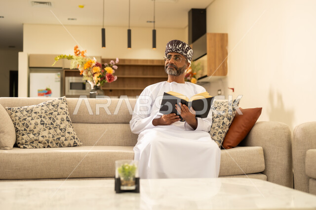 مفهوم الشغف بالقراءة, إيماءات التركيز والاندماج ، ثقافة التعلم الذاتي، قراءة الكتب بتمعن ، رجل عربي خليجي عماني يرتدي الدشداشة والكمة يجلس على اريكة مريحة ويحمل بيده كتاب تثقيفي ،  النظر  بعيداً الى مكان ما ، الاستمتاع بقراءة الروايات والقصص .