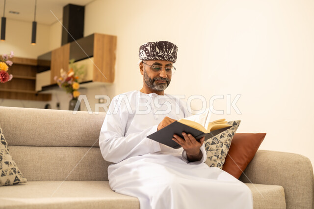 Self-learning culture, reading books carefully, an Arab Gulf Omani man wearing a dishdasha and kimmah sitting on a comfortable sofa holding an educational book, gestures of concentration and engagement, enjoying reading novels and stories.
