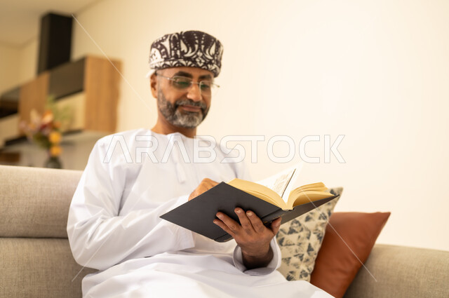 القراءة بإيماءات التركيز والاندماج, الشغف بممارسة الهوايات المفيدة ، صورة مقربة لرجل عربي خليجي عماني يرتدي الدشداشة والكمة يجلس على اريكة مريحة ويحمل بيده كتاب تثقيفي ، قراءة الكتب بتمعن ، الاستمتاع بقراءة الروايات والقصص .