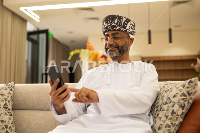 النقر بإصبع السبابة على شاشة الجوال ، الدردشة مع الأهل والأصدقاء عبر الانترنت، استخدام التكنولوجيا و التقنيات الحديثة, صورة مقربة لرجل عربي خليجي يرتدي الدشداشة والكمة ويحمل بيده الهاتف المحمول ، تصفح مواقع و وسائل التواصل الاجتماعي .